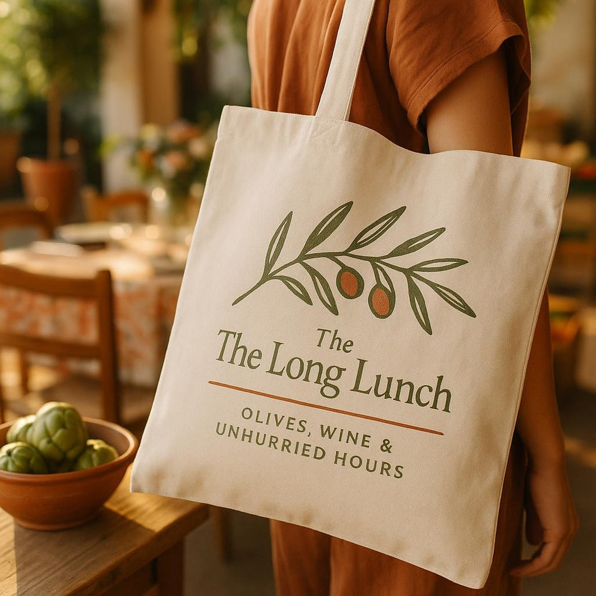 The canvas tote resting on a sunlit Mediterranean table beside olives and a linen napkin