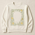 Cream unisex sweatshirt with hand-drawn lemon and olive branch border design, shown flat on a linen backdrop