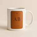 Tan and cream ceramic mug printed with a vegetable-tanned leather luggage tag illustration on a white background